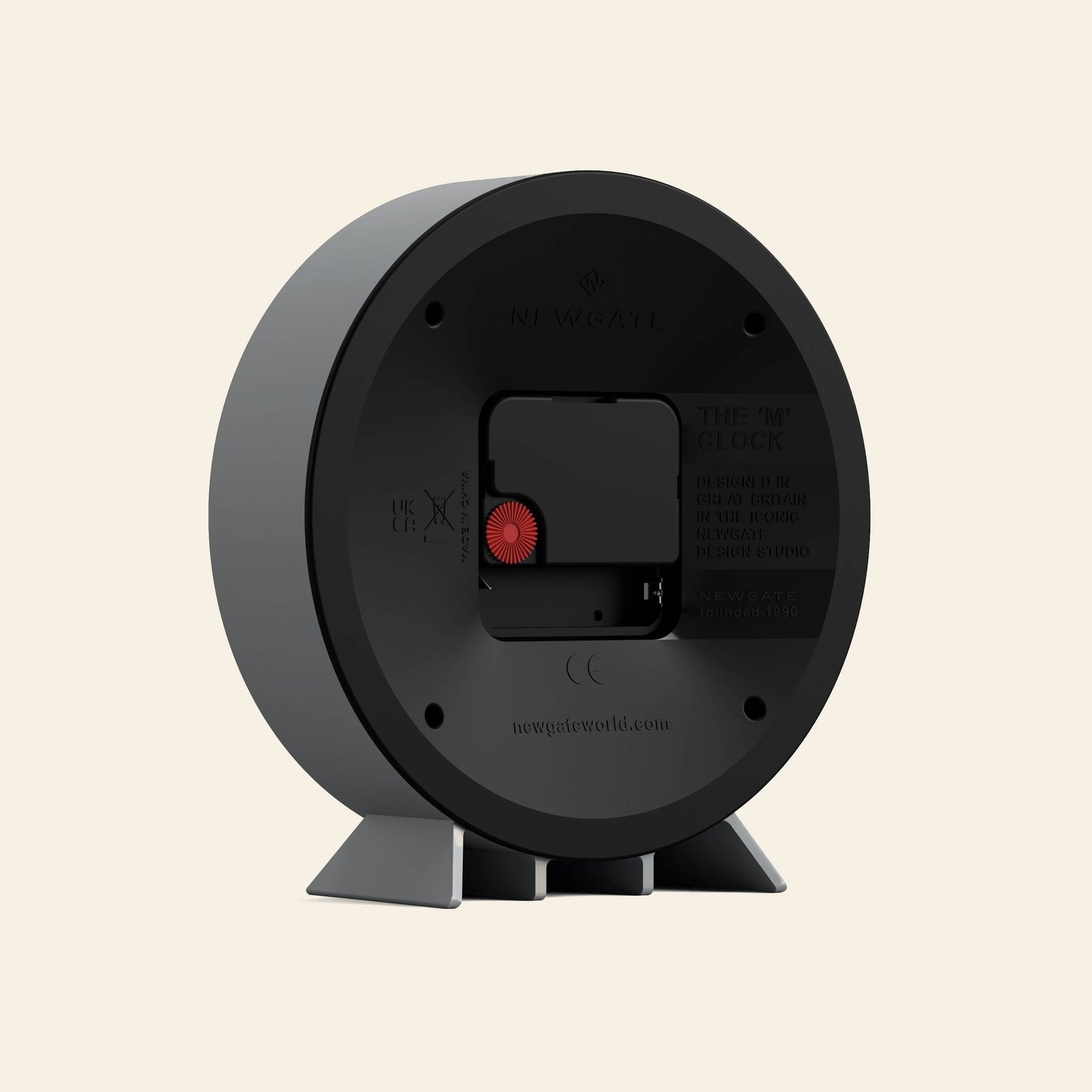 The image shows the back of the Newgate M Mantel Railway Clock, featuring a battery compartment, adjustment dial, two gray support legs, classic design, and Newgate branding with product details on the back.