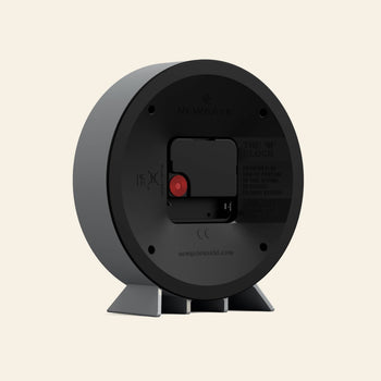 The image shows the back of the Newgate M Mantel Railway Clock, featuring a battery compartment, adjustment dial, two gray support legs, classic design, and Newgate branding with product details on the back.