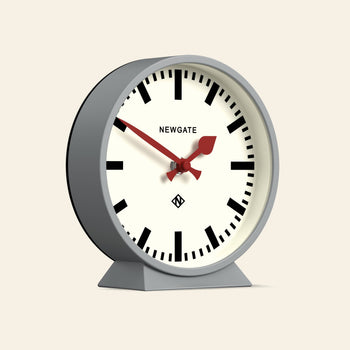 The Newgate M Mantel Railway Clock features a white face, bold black hour marks, red hands, and a gray body on a stand. Newgate is printed on the face, showing the time as 9:22—echoing classic railway clock design.