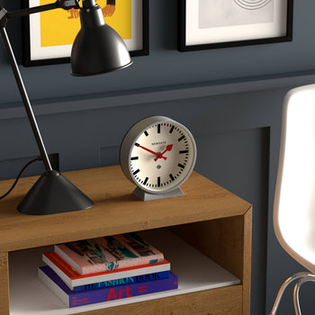A modern desk with a black lamp, a small stack of colorful books, and the Newgate M Mantel Railway Clock by Newgate showing 10:11. Behind, a dark wall displays framed art with part of a white chair visible.