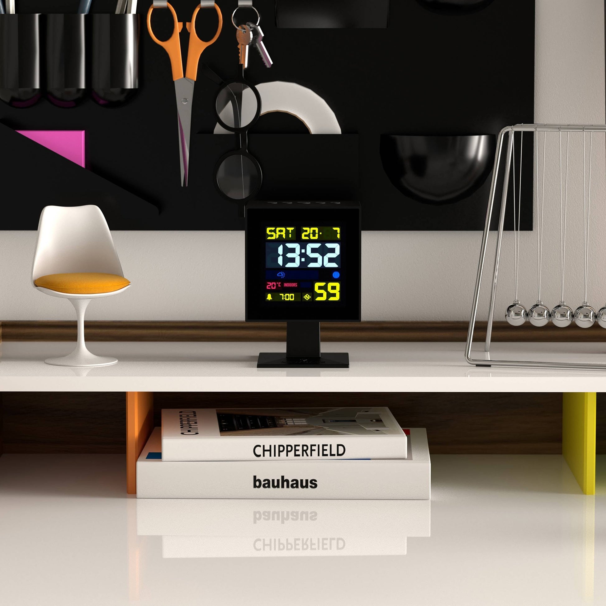 A modern desk displays the Newgate Monolith LCD Alarm Clock by Newgate showing time, weather, and moon phase, alongside a Newton’s cradle, a white lamp, orange and white books, scissors, and kitchen utensils on a black wall organizer.