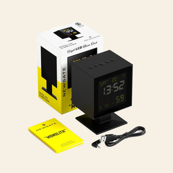 A black Newgate Monolith LCD Alarm Clock with a square display sits on a stand. Next to it are a USB charging cable, yellow instructions, and its white and yellow Newgate packaging featuring moon phase details.
