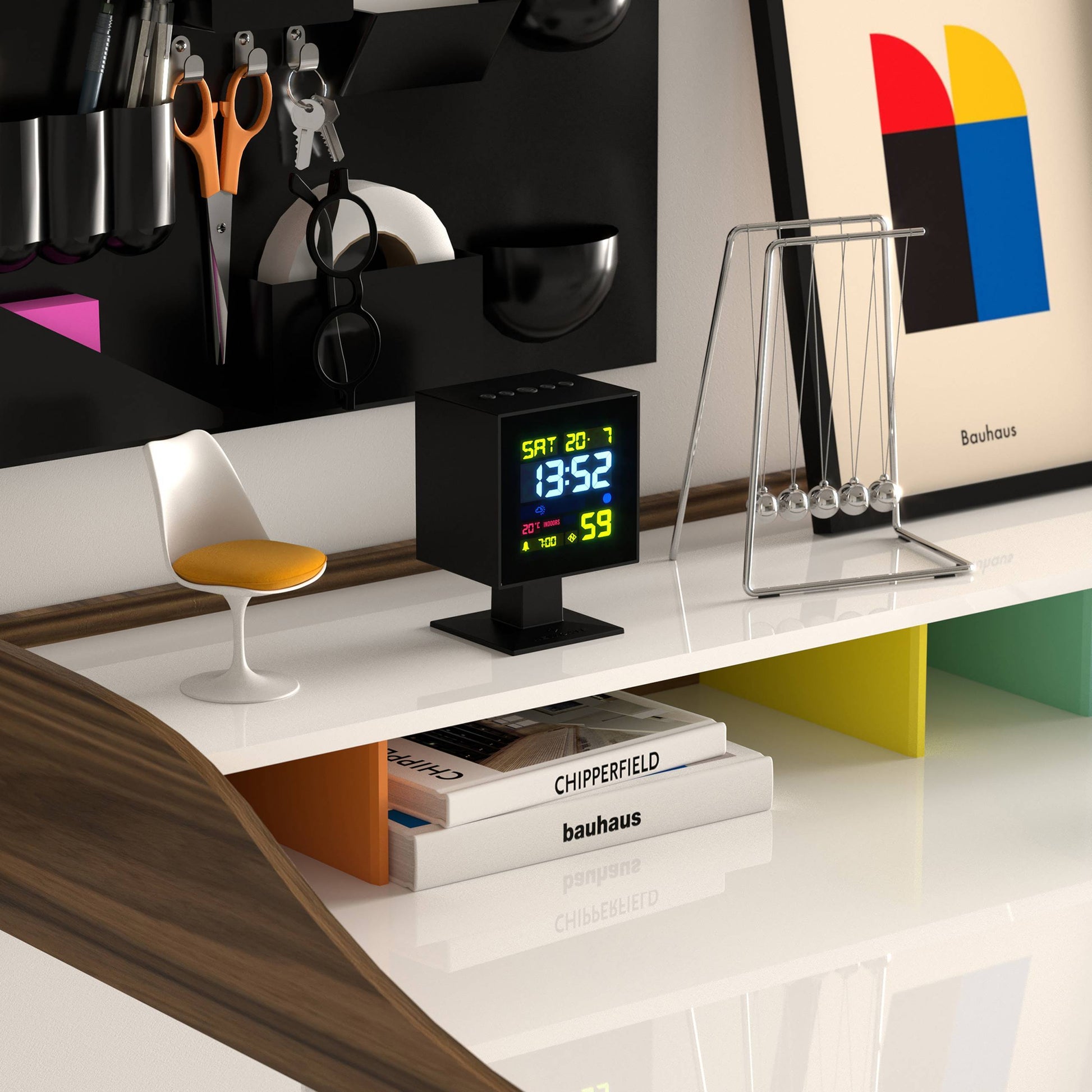A sleek modern desk with a Newgate Monolith LCD Alarm Clock displaying moon phase and weather, Newton’s cradle, a white-and-yellow chair, scissors, colorful Bauhaus artwork, and “Chipperfield” books on a white shelf.