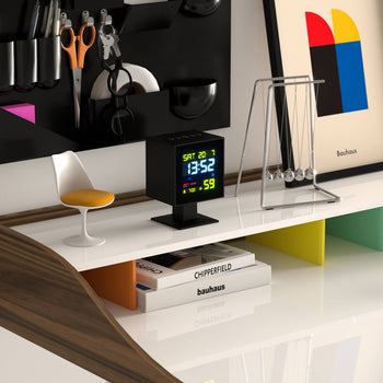 A sleek modern desk with a Newgate Monolith LCD Alarm Clock displaying moon phase and weather, Newton’s cradle, a white-and-yellow chair, scissors, colorful Bauhaus artwork, and “Chipperfield” books on a white shelf.