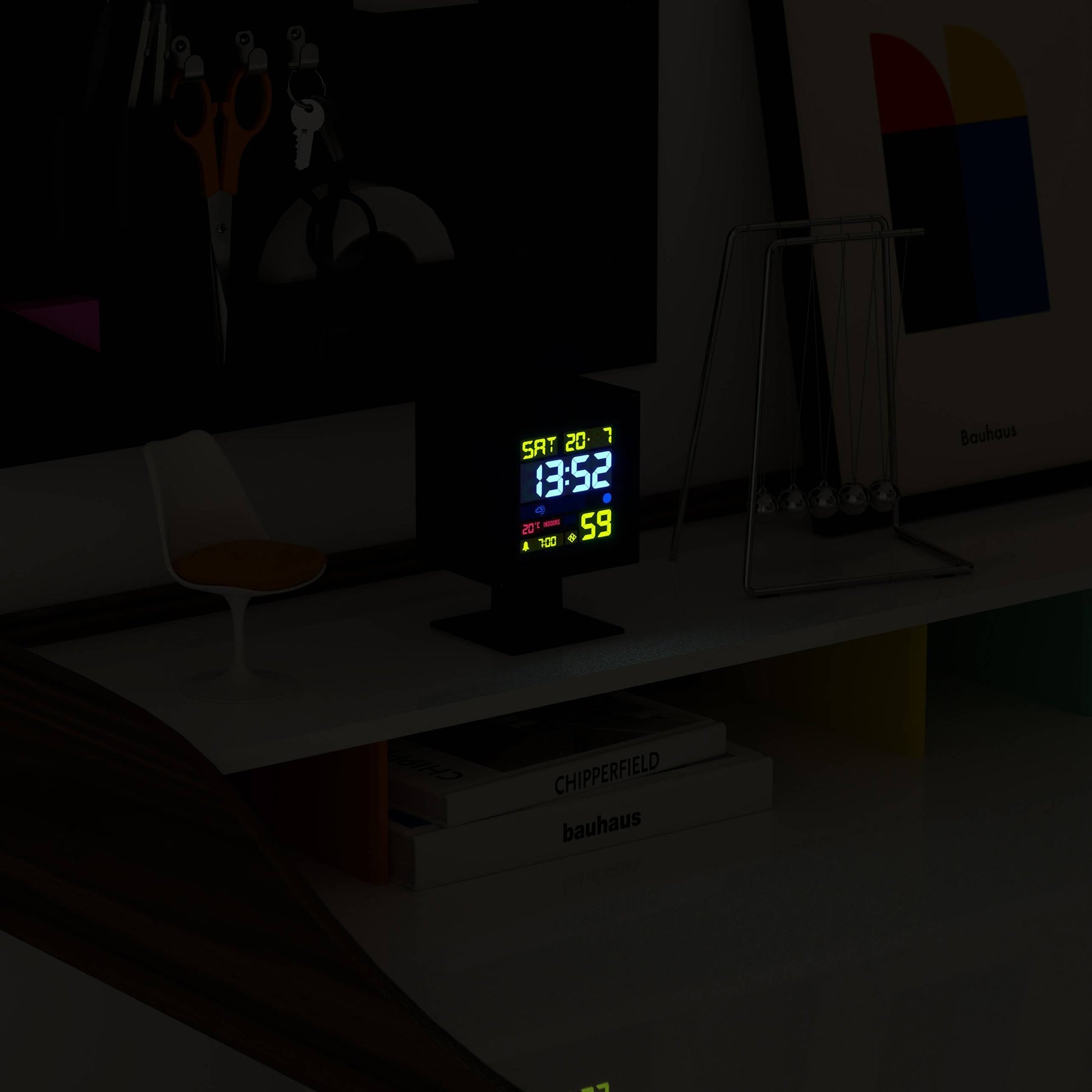 The Newgate Monolith LCD Alarm Clock by Newgate, showing 13:52, Sat 20.1, and a moon phase icon, sits on a minimalist white shelf with a Newtons cradle, framed art, chair, and books for a modern touch.