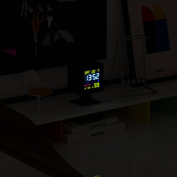 The Newgate Monolith LCD Alarm Clock by Newgate, showing 13:52, Sat 20.1, and a moon phase icon, sits on a minimalist white shelf with a Newtons cradle, framed art, chair, and books for a modern touch.