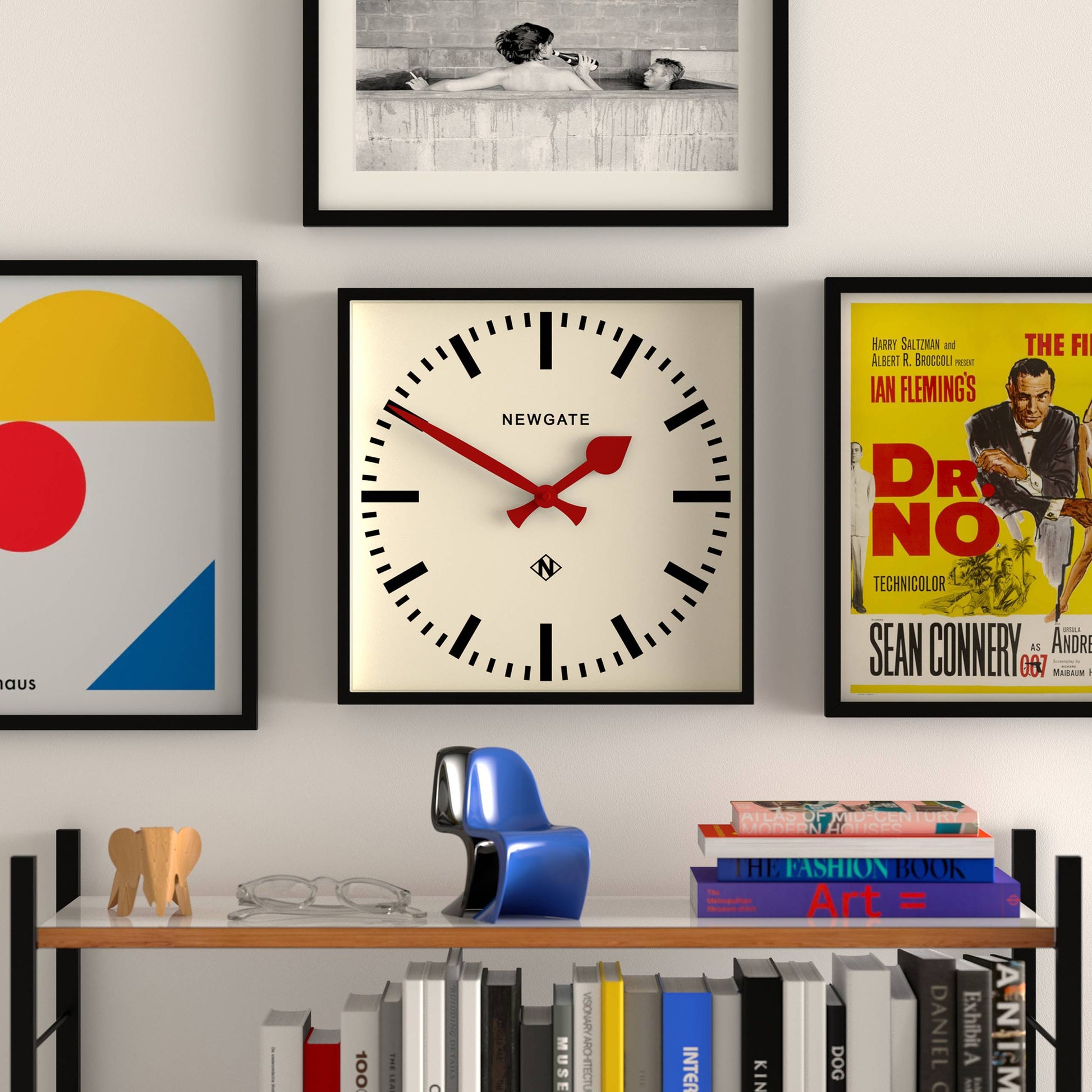 The Newgate Number Five Railway Wall Clock by Newgate, featuring vibrant red hands and a minimalist black case, hangs on a white wall amid framed art. Below, books, glasses, a blue decor item, and a wooden elephant rest on a shelf.