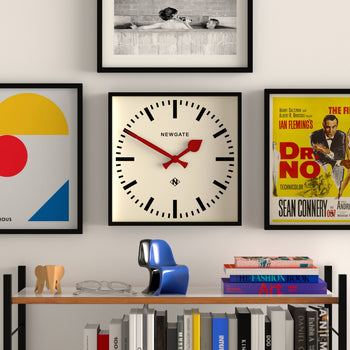 The Newgate Number Five Railway Wall Clock by Newgate, featuring vibrant red hands and a minimalist black case, hangs on a white wall amid framed art. Below, books, glasses, a blue decor item, and a wooden elephant rest on a shelf.