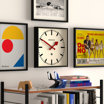 The Newgate Number Five Railway Wall Clock by Newgate, featuring vibrant red hands and a minimalist black case, is mounted among framed art prints above a bookshelf showcasing books, a small sculpture, and decorative items.