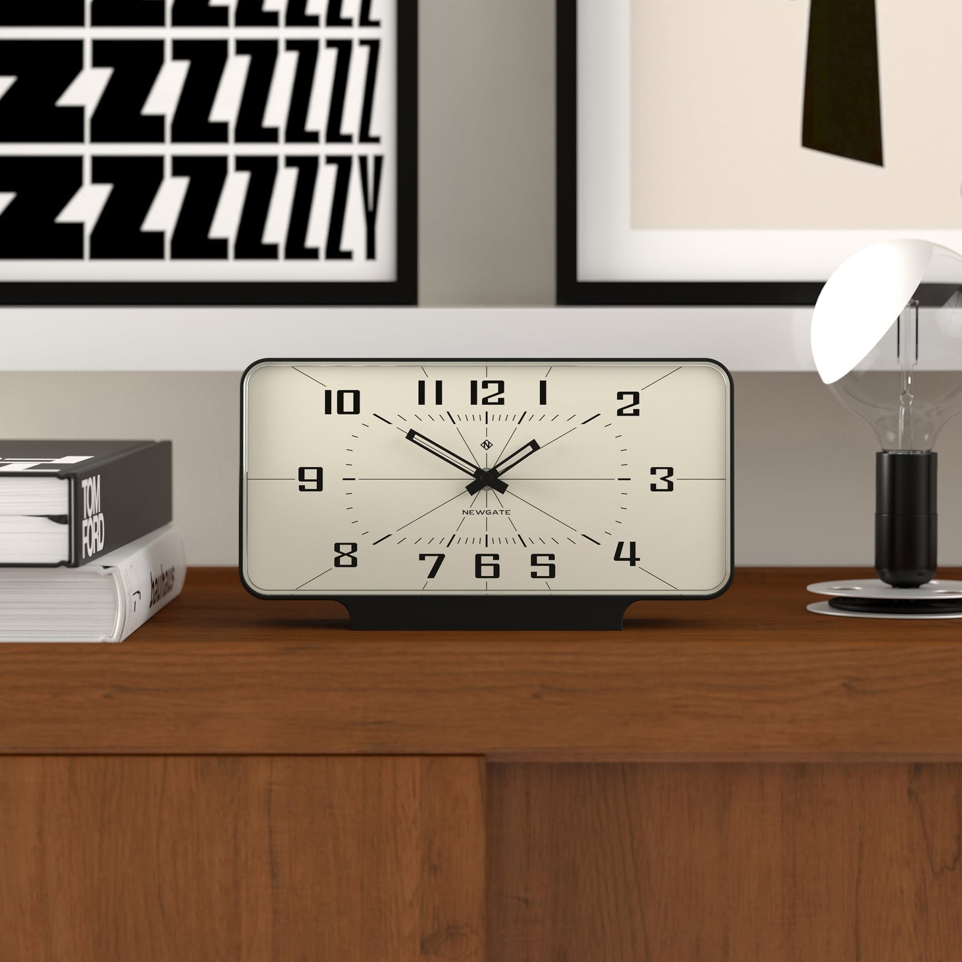 The Newgate Planet Mantel Clock by Newgate, featuring Arabic numerals, sits on a wooden surface beside books and a lamp, with framed artwork on the wall behind it. The clock displays the time as 10:09:34.