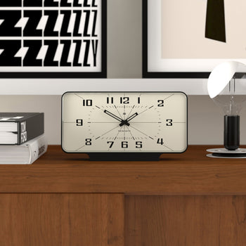 The Newgate Planet Mantel Clock by Newgate, featuring Arabic numerals, sits on a wooden surface beside books and a lamp, with framed artwork on the wall behind it. The clock displays the time as 10:09:34.