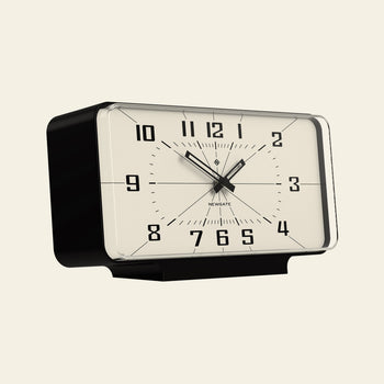 The Newgate Planet Mantel Clock by Newgate is a rectangular, vintage-style black and white clock with bold Arabic numerals and hands, shown from a slight left angle against an off-white background.