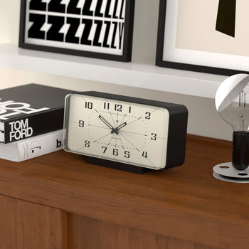 The Newgate Planet Mantel Clock by Newgate, in black and white with Arabic numerals, sits on a wooden shelf set to 11:11. Nearby are a Tom Ford book, a black and white framed print, and a decorative light bulb.