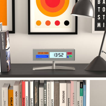 The Newgate Supergenius LCD Alarm Clock by Newgate displays 13:52 and temperature as it rests on a dark shelf beside a black desk lamp, books, and vibrant artwork; its USB cable keeps it connected for convenience.
