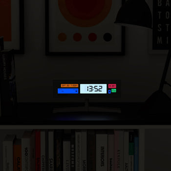 A Newgate Supergenius LCD Alarm Clock by Newgate, showing 13:52, rests on a dark shelf among books and glows under a desk lamp. Framed artwork hangs behind it, while a USB cable powers the clock.