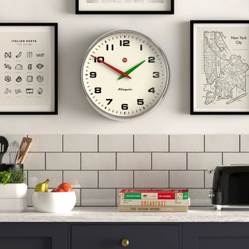A Newgate Superstore Wall Clock by Newgate shows 10:10 above a countertop with a bowl of fruit, kitchen utensils, stacked recipe books, and a black toaster, set against framed art and a white-tiled backsplash.