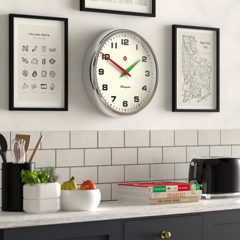 A kitchen wall features a Newgate Superstore Wall Clock showing 12:03, framed art prints, a countertop with fruit, utensils, a toaster, jars, stacked cookbooks, and a white subway tile backsplash.