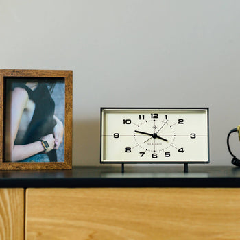 A framed photo of a woman, the Newgate Wideboy Alarm Clock by Newgate showing 10:11 with a mid-century atomic dial, and a coiled cord sit on a wooden surface against a light gray wall.