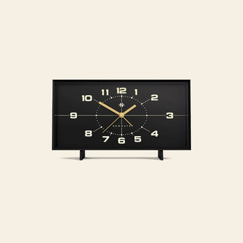 The Newgate Wideboy Alarm Clock by Newgate is a rectangular black clock with gold hands and numbers, a minimalist mid-century atomic dial, and two small legs, shown against a plain beige background.