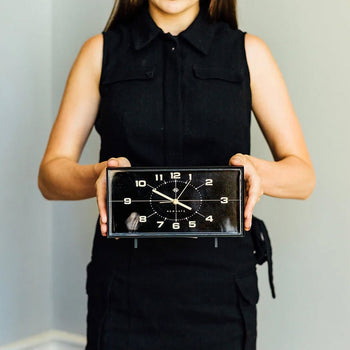 A woman in a sleeveless black dress holds the Newgate Wideboy Alarm Clock by Newgate, featuring a rectangular retro design with white numbers and hands, standing against a plain light background.