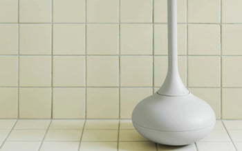 The Normann Copenhagen Ballo Toilet Brush, in white, features a rounded base and stands on a tiled bathroom floor beside a wall of square pale yellow tiles.