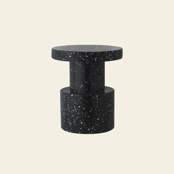The Normann Copenhagen Bit Stool, a compact modern black seat with white speckles, features a round top and cylindrical base, highlighting its multifunctional design against a plain off-white background.