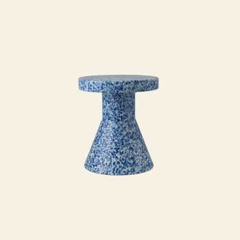 The Normann Copenhagen Bit Stool Cone features a small, round silhouette with a flared base, speckled in blue and white hues. Its multifunctional design works as a side table or extra seating, shown against a plain cream background.
