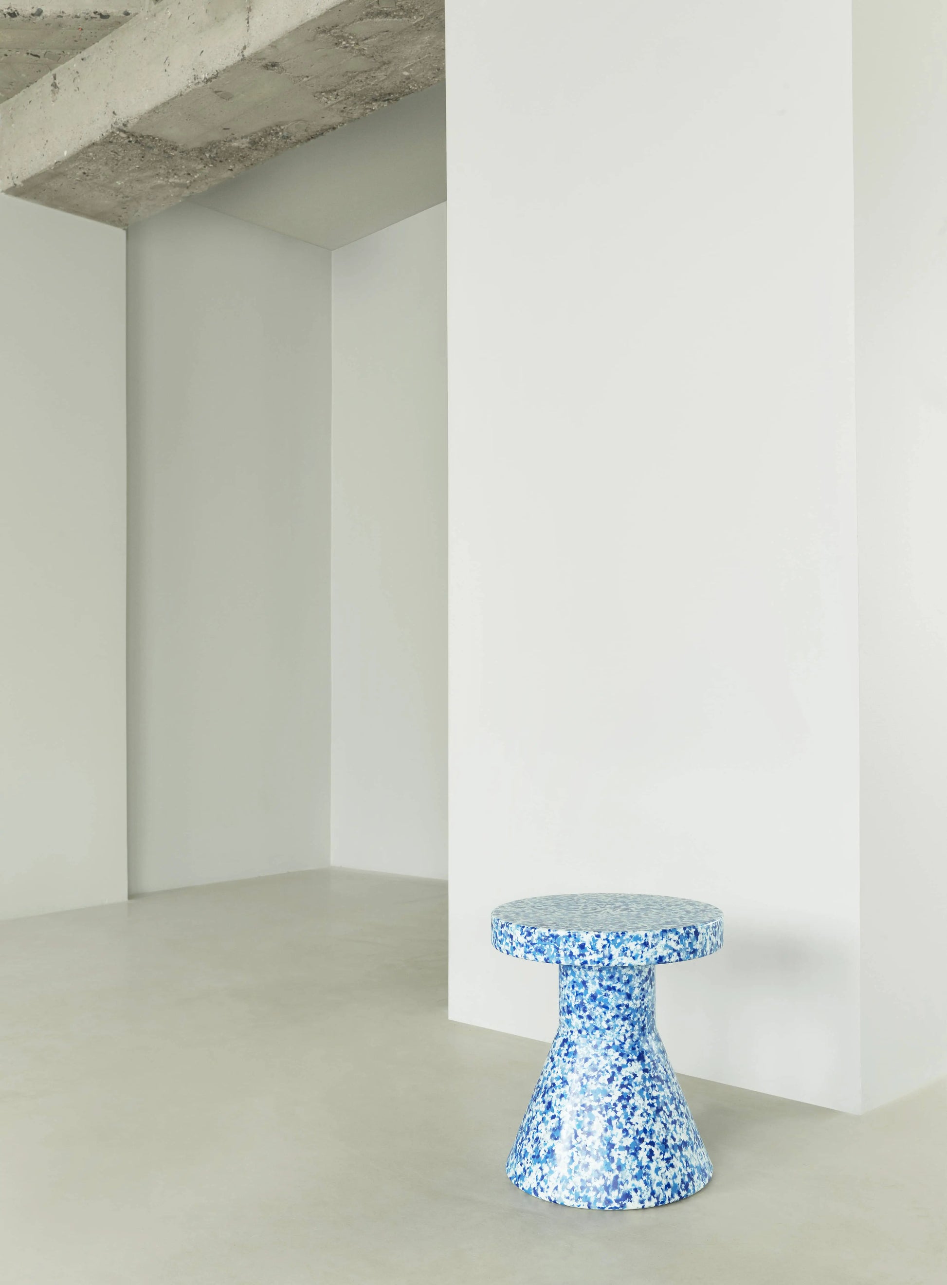 A compact, blue and white speckled Normann Copenhagen Bit Stool Cone from Normann Copenhagen adds multifunctional style to a minimalist room with white walls, a concrete ceiling, and a pale floor.