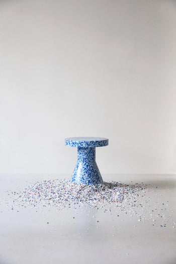 A Normann Copenhagen Bit Stool Cone stands in the center of a light gray room, its blue speckled design and multifunctional use showcased by colorful confetti-like pieces scattered at its base.