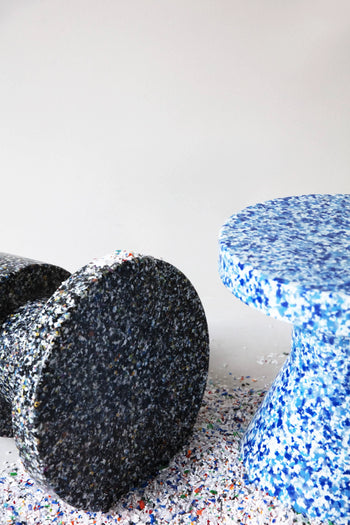 Two Normann Copenhagen Bit Stool Cone seats made from recycled plastic are displayed on a plain background—one upright in blue with white speckles, the other on its side in black with colorful flecks. Plastic fragments surround them.