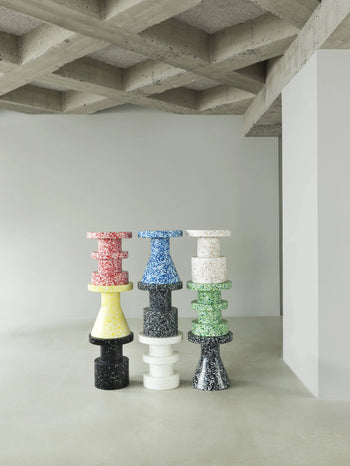 Four colorful Normann Copenhagen Bit Stool Stack pieces are arranged in a minimalist room with concrete floors and a coffered ceiling. Each stool features stacked, abstract shapes for versatile seating and multifunctional design.