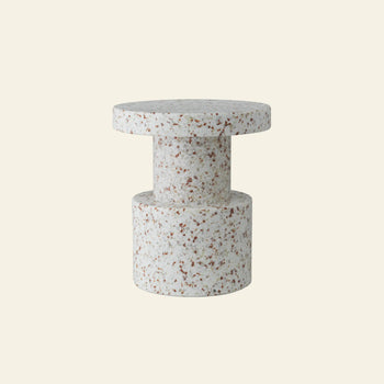 The Normann Copenhagen Bit Stool by Normann Copenhagen is a modern cylindrical side table in speckled white and brown terrazzo, with a multifunctional design featuring a thick round top and wide base, shown against a plain beige background.