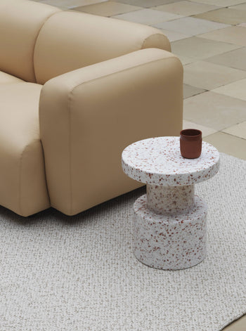 A beige sofa sits on a textured light rug beside a round, speckled terrazzo side table and the Normann Copenhagen Bit Stool, offering versatile seating in a space with neutral-toned tiled floors.