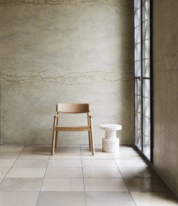 A wooden chair with a tan seat and backrest sits beside a Normann Copenhagen Bit Stool near a large window in a minimalist room with tiled floors and a textured beige wall, highlighting versatile seating by Normann Copenhagen.
