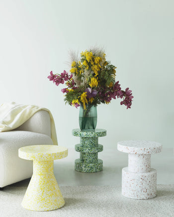 A modern living space displays a white textured chair, cream blanket, and three Normann Copenhagen Bit Stools in yellow, green, and white as side tables, plus a green vase with vivid yellow and purple flowers—highlighting versatile design.