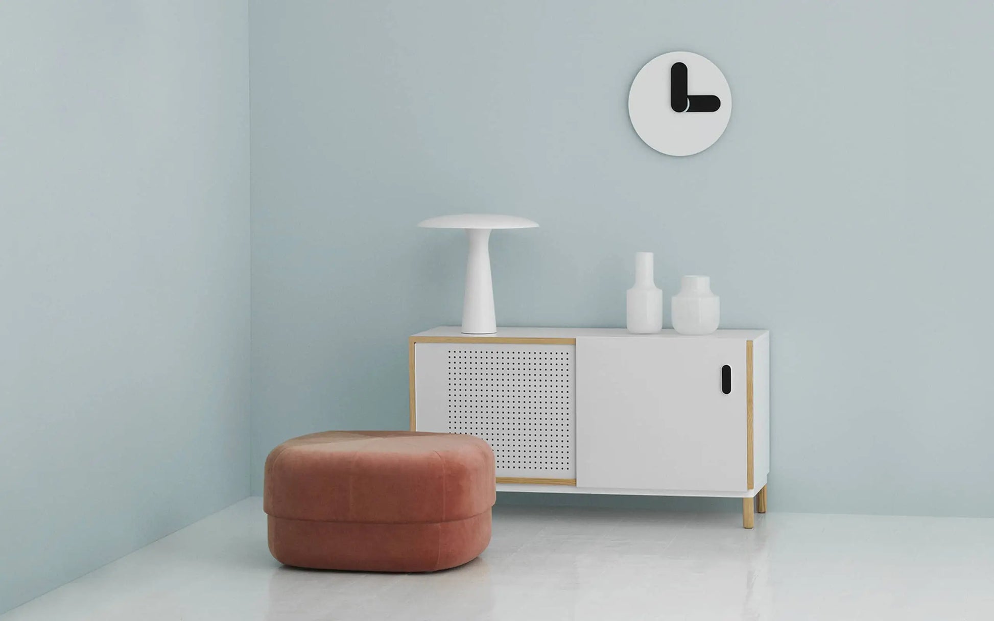 A minimalist room with Danish flair: a white cabinet, two white vases, and a white lamp below the Normann Copenhagen Bold Wall Clock. A round brown ottoman sits on the white floor against a light blue wall.
