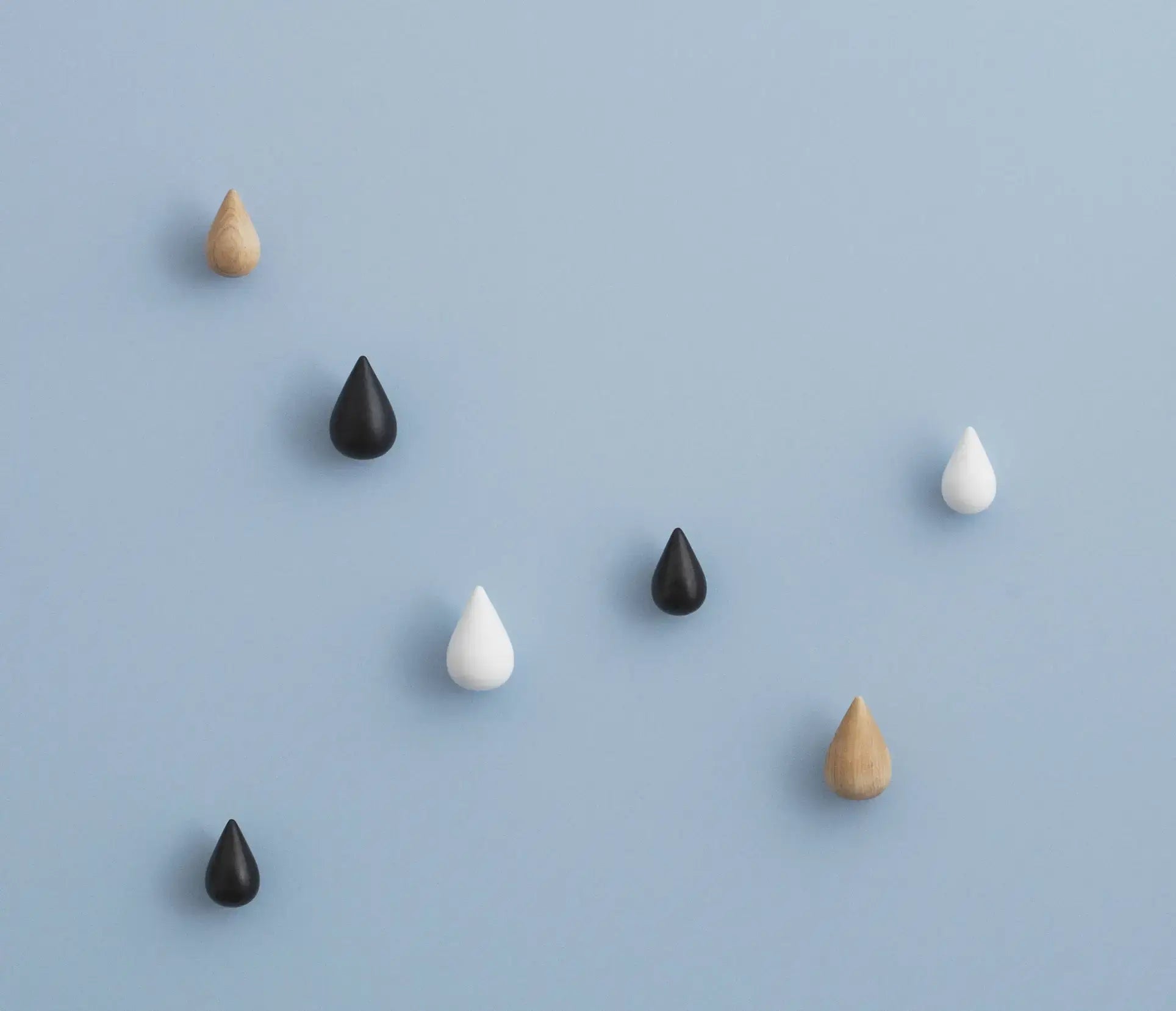 Seven Normann Copenhagen Dropit Hooks Large, Set of 2 by Normann Copenhagen in black, white, and wood hues are scattered on a light blue surface, forming an eye-catching irregular wooden pattern.