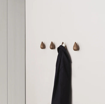 A clean, modern wall features Normann Copenhagen Dropit Hooks Large (set of 2) in wood. One teardrop-shaped hook holds a dark coat, while the other remains empty, showcasing their minimalist Danish design.