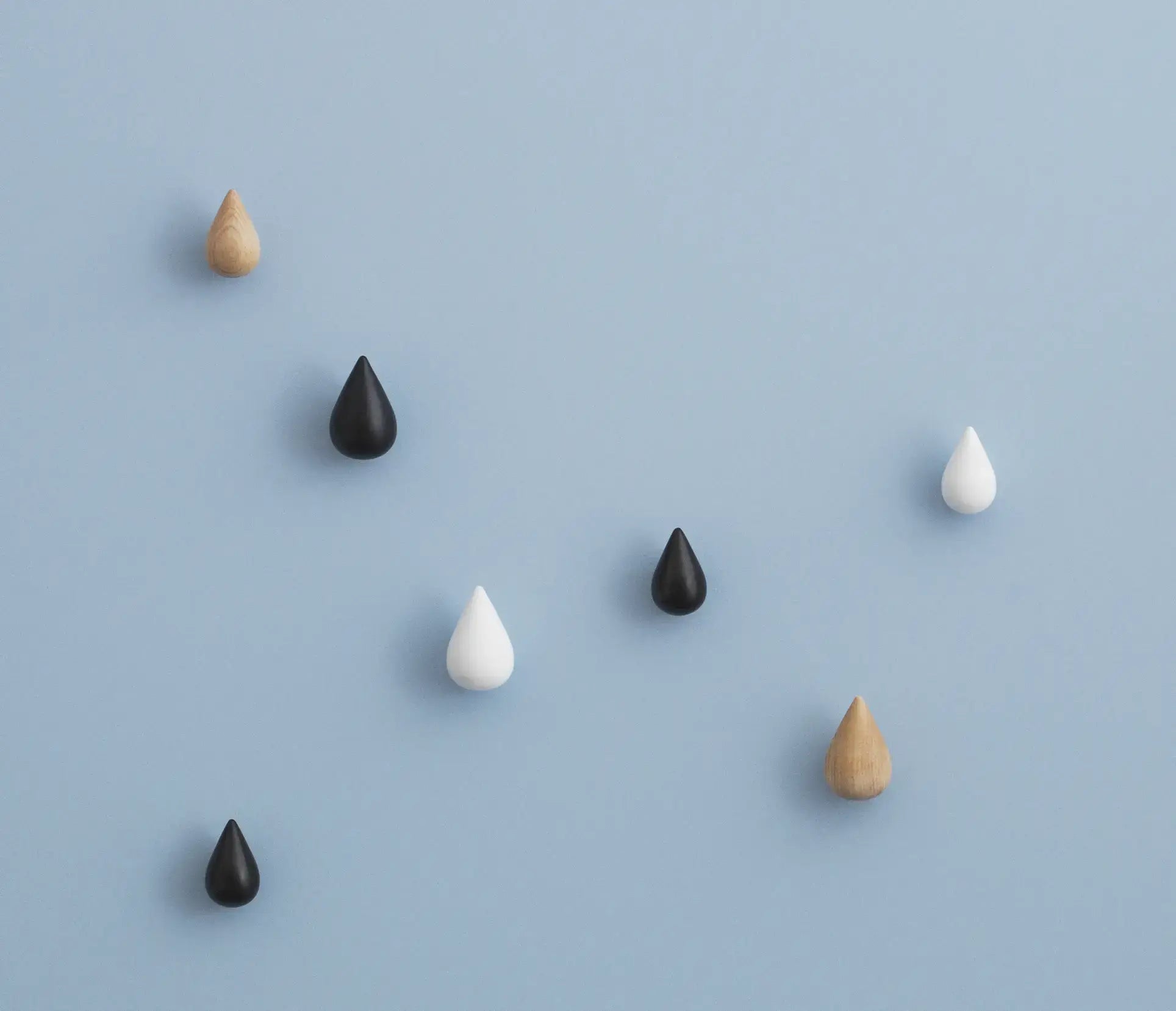 Seven Normann Copenhagen Dropit Hooks Small, Set of 2 by Normann Copenhagen, in black, white, oak, and dark walnut finishes, are scattered on a light blue background.
