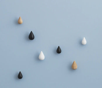 Seven Normann Copenhagen Dropit Hooks Small, Set of 2 by Normann Copenhagen, in black, white, oak, and dark walnut finishes, are scattered on a light blue background.