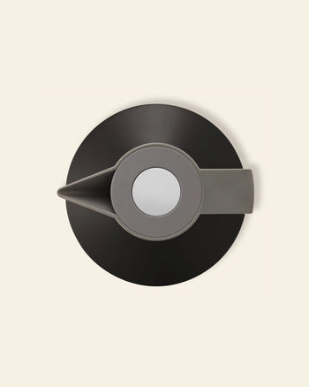 The Normann Copenhagen Geo Vacuum Jug, in grey and black, is seen from above with its round lid, spout, and handle. Its vacuum-insulated body by Normann Copenhagen keeps drinks hot or cold for hours against a beige background.