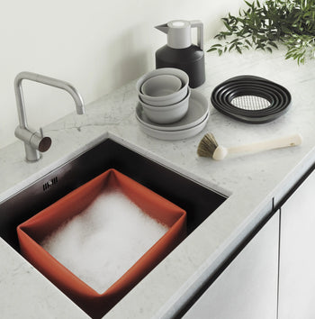 A modern kitchen sink with an orange wash basin, soapy water, stacked dishes, a scrubbing brush, black plates, green leaves, and the Normann Copenhagen Geo Vacuum Jug by Normann Copenhagen on a white marble countertop.