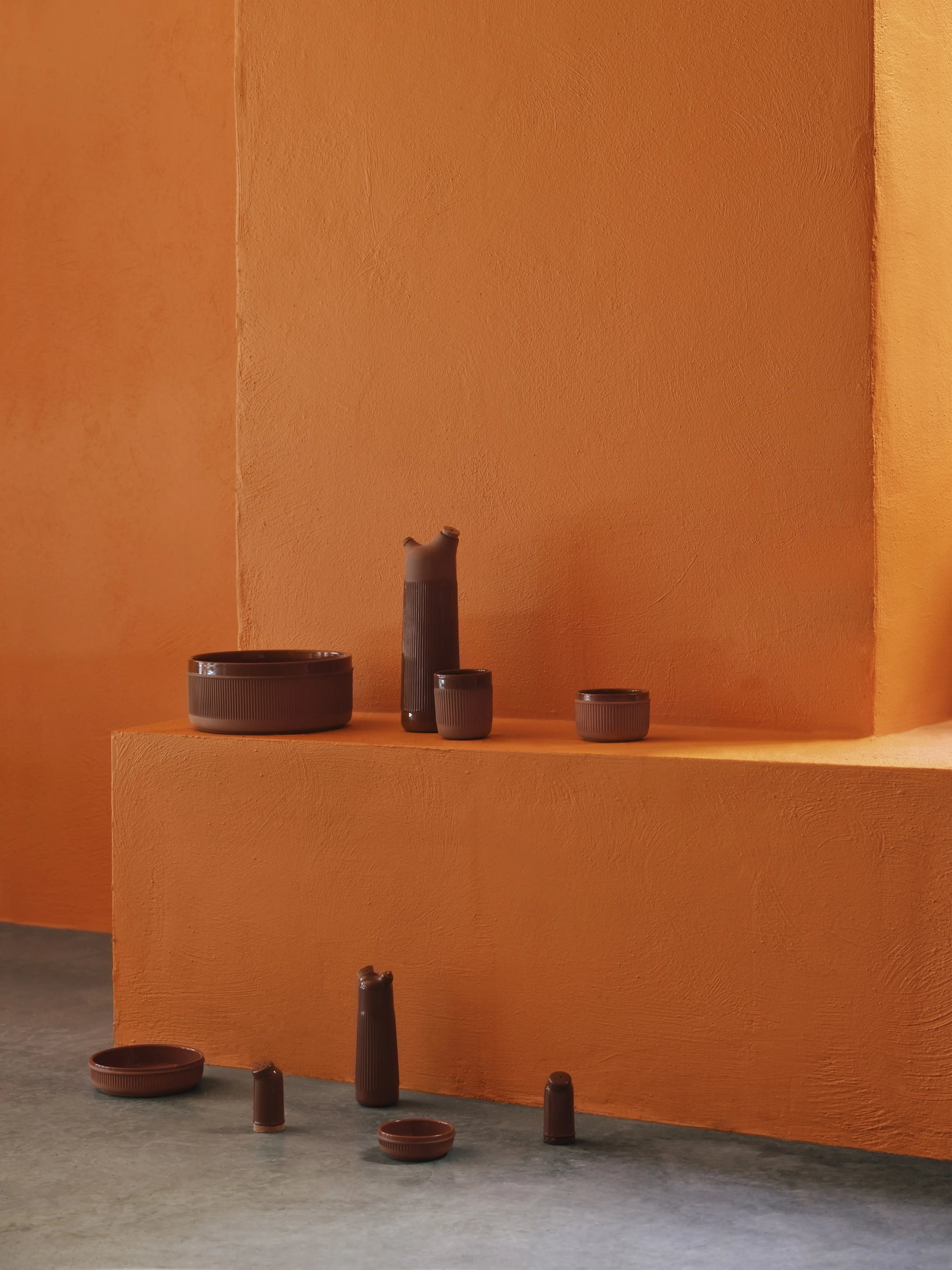 Rustic terracotta vessels, including the 15cm Normann Copenhagen Junto Bowl, are arranged on and before an orange textured platform set against an orange wall and concrete floor.