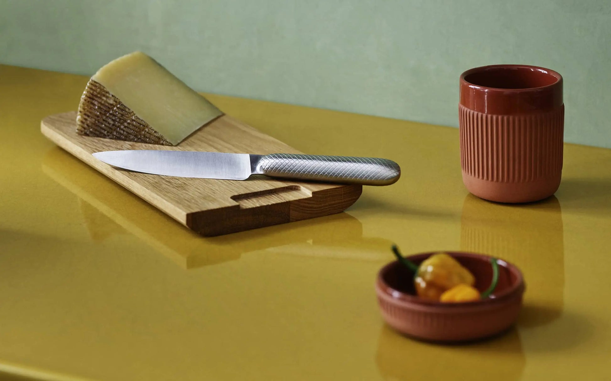 A knife sits on a wooden cutting board with cheese, beside a red Normann Copenhagen mug and the versatile Normann Copenhagen Junto Dish (15.5cm) in terracotta holding yellow peppers, all atop a glossy yellow surface.