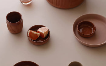 A minimalist table setting showcases Normann Copenhagen Junto Dish, 15.5cm, with earthy ceramic tones, grapefruit wedges, bowls, cups, and plates artfully arranged on a light surface.