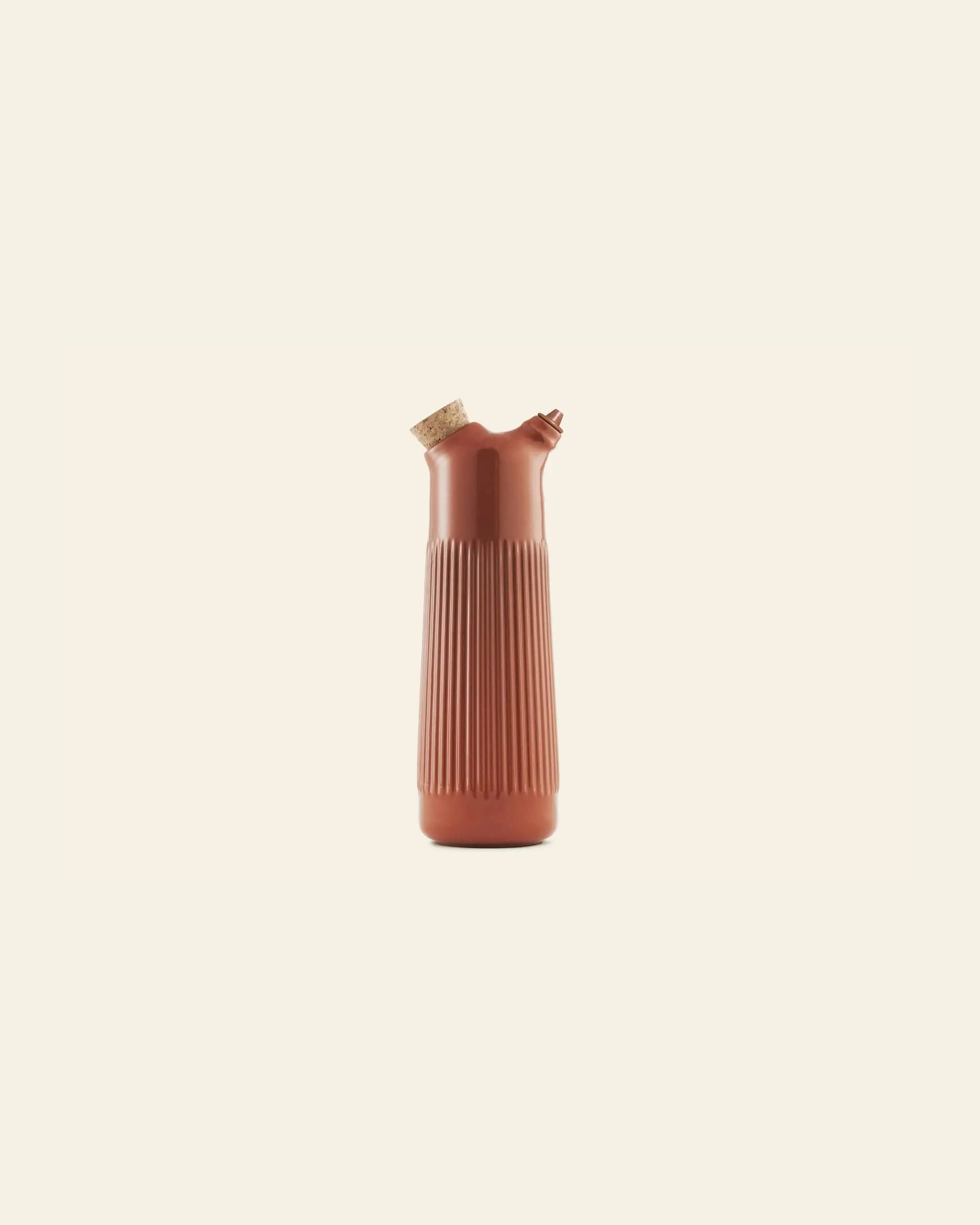 The Normann Copenhagen Junto Oil Bottle by Normann Copenhagen, featuring vertical ridges, a cork stopper, and a small spout in reddish-brown earthenware, stands upright against a plain light beige background.