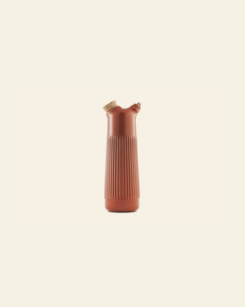 The Normann Copenhagen Junto Oil Bottle by Normann Copenhagen, featuring vertical ridges, a cork stopper, and a small spout in reddish-brown earthenware, stands upright against a plain light beige background.