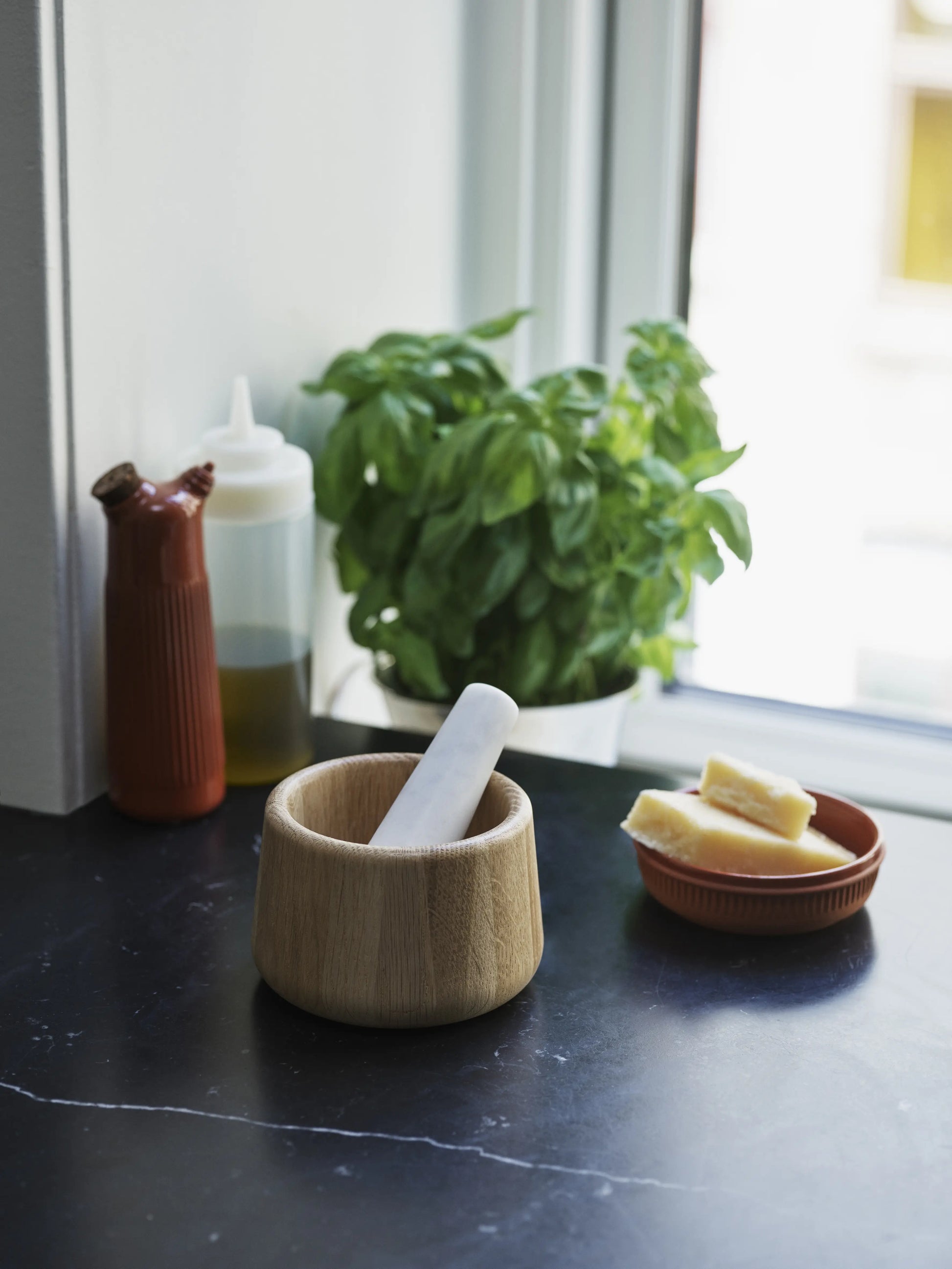 A wooden mortar with a white pestle sits on a dark countertop beside cheese, basil, and the Normann Copenhagen Junto Oil Bottle—an inviting scene of cozy charm inspired by Normann Copenhagen.