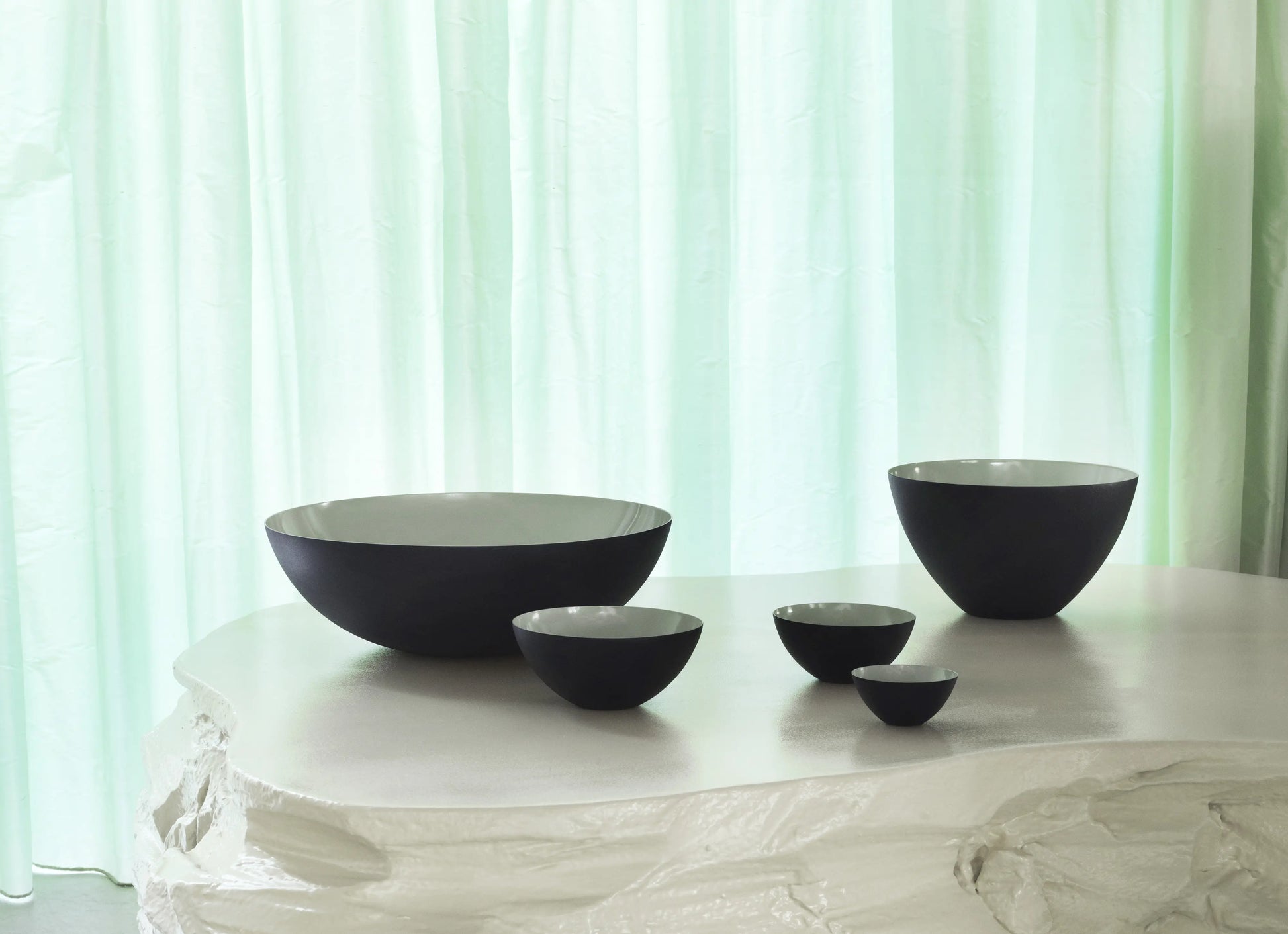 A set of five Normann Copenhagen Krenit Bowls, 12.5cm, in black and white, are arranged on a textured white surface with a pale green curtain backdrop, reflecting iconic Danish design.