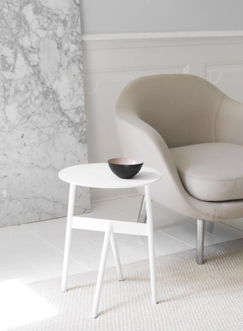 A modern beige armchair sits beside a small round white side table topped with the 12.5cm Krenit Bowl by Normann Copenhagen. A marble slab and white walls create a minimalist, elegant Danish design backdrop.